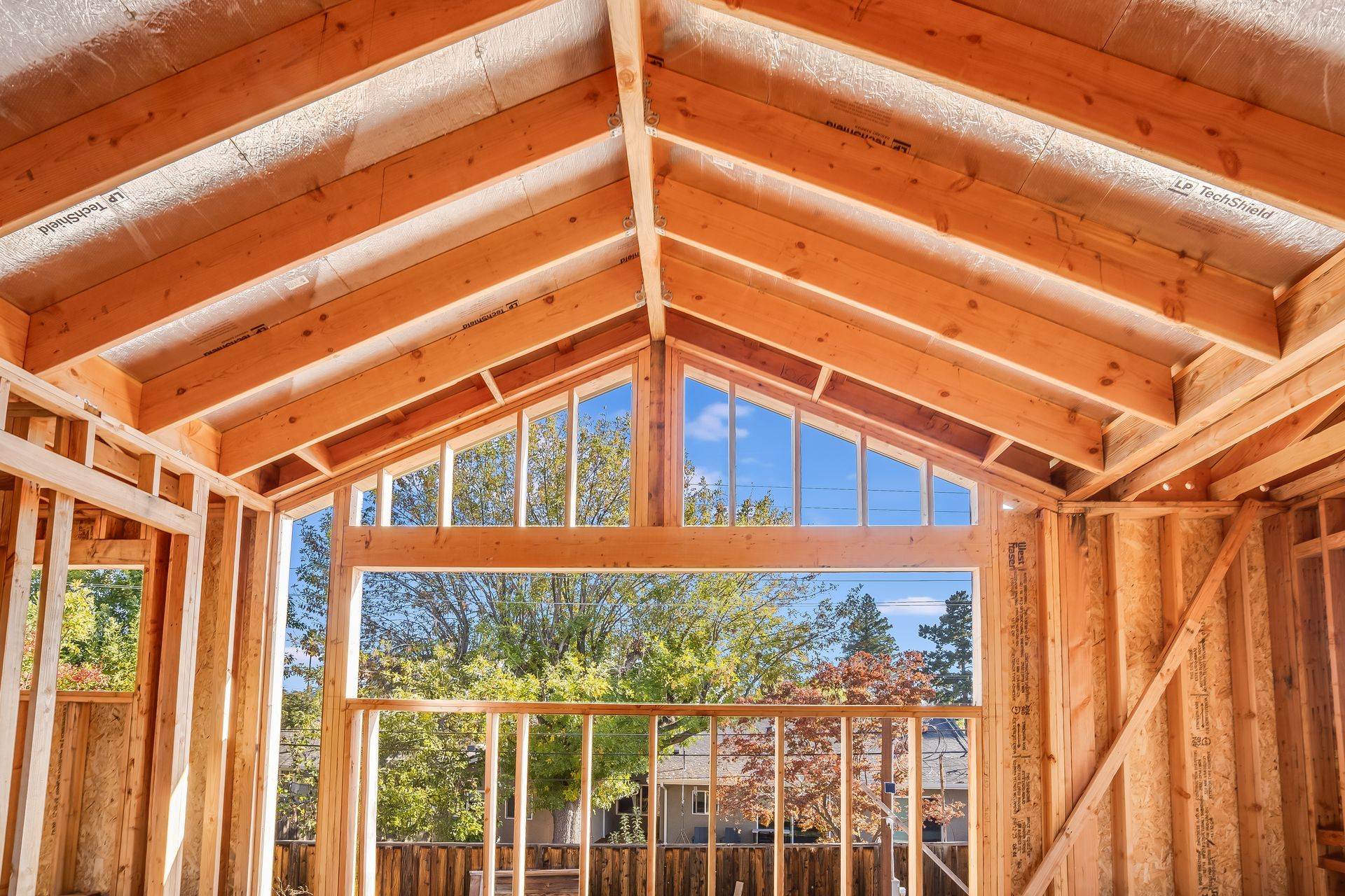 Interior view of a building frame with exposed wooden beams, open windows, and a view of trees and sky.