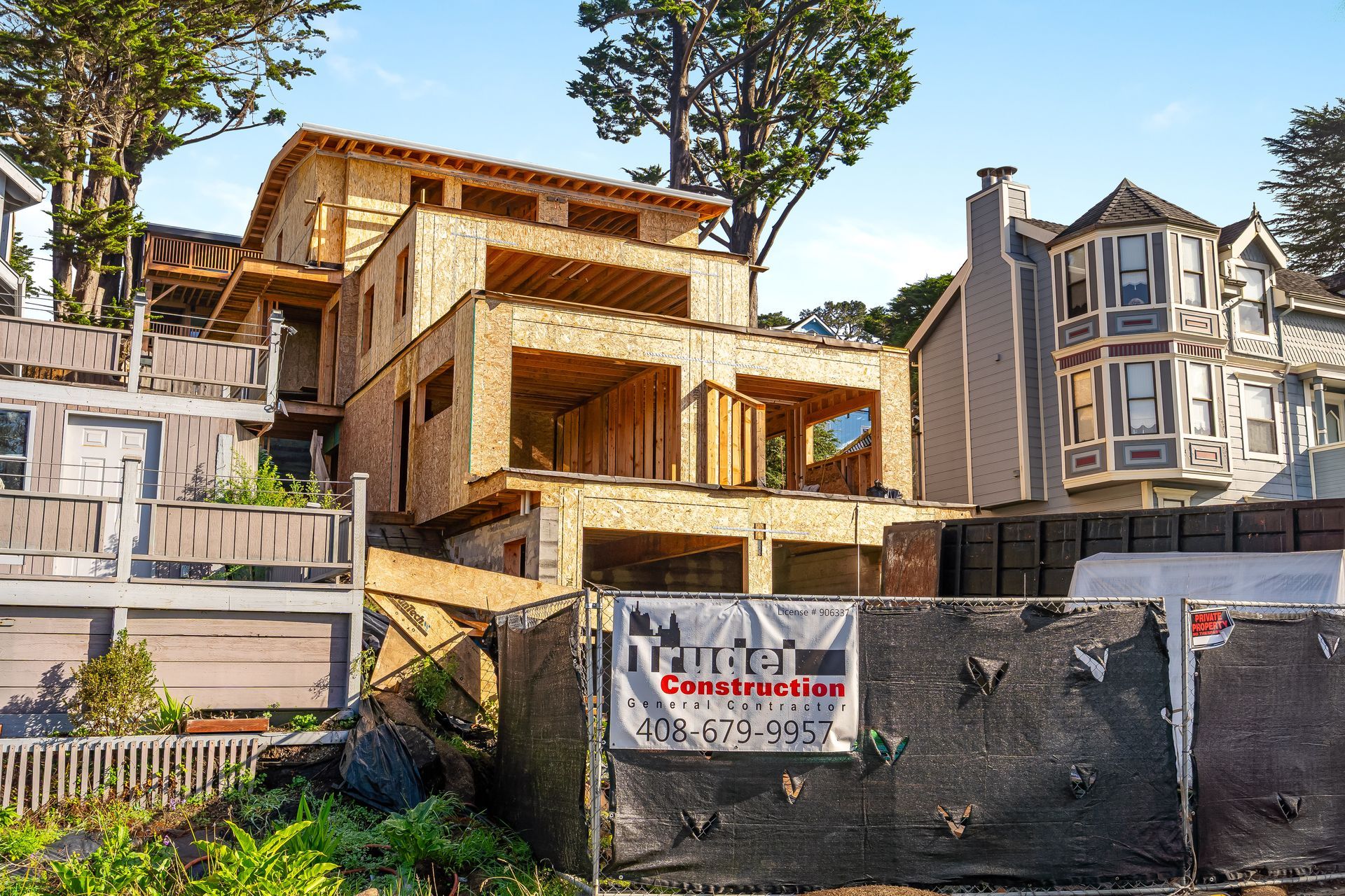 Construction of a multi-story house, wooden frame visible. Fence in front with 