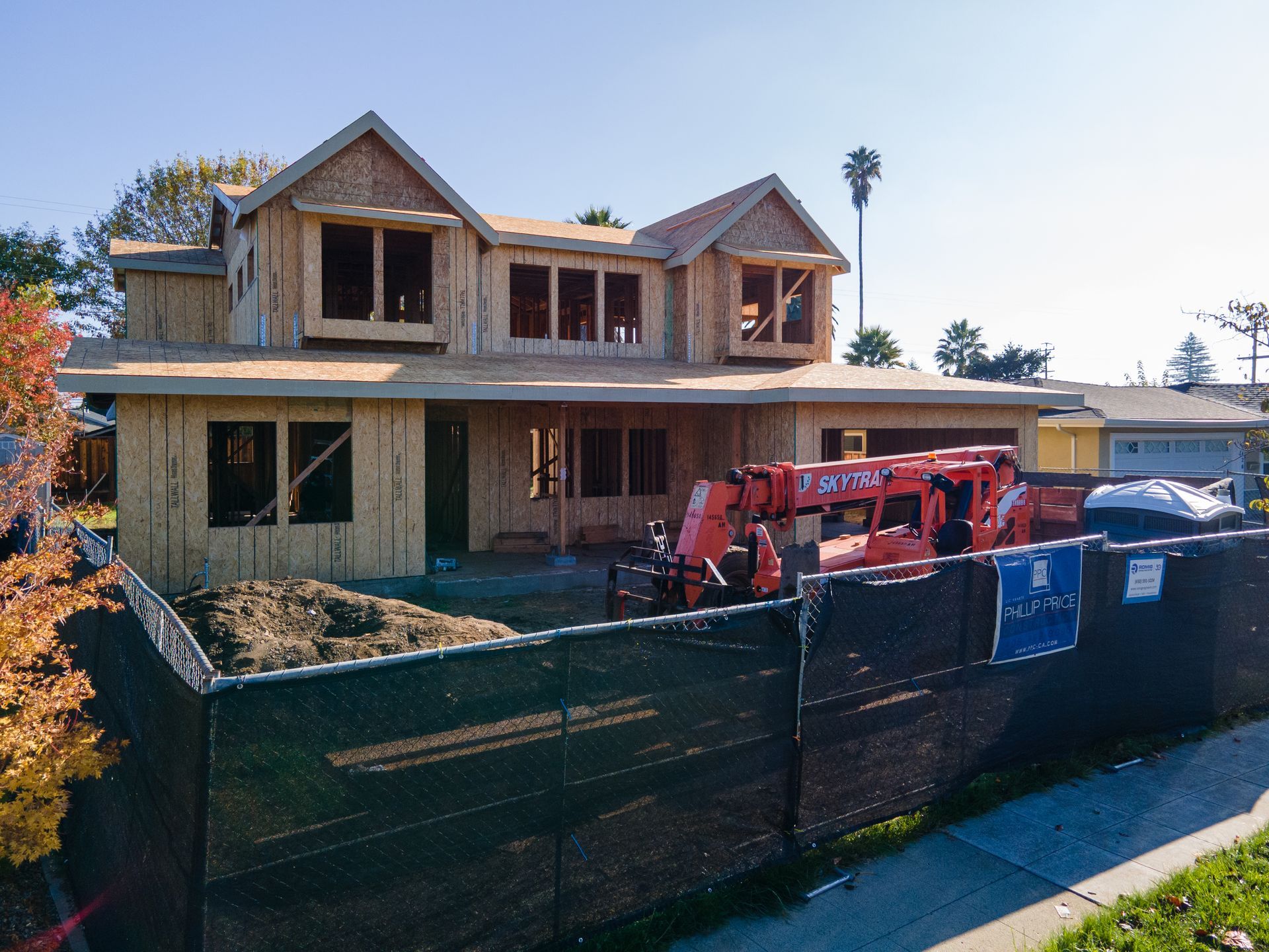 Two-story house under construction with exposed wood framing, orange crane, and black fencing in a residential area.