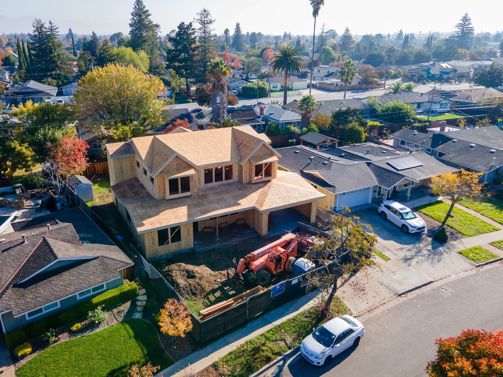 Aerial view of a house under construction in a suburban neighborhood, framed by trees and houses.