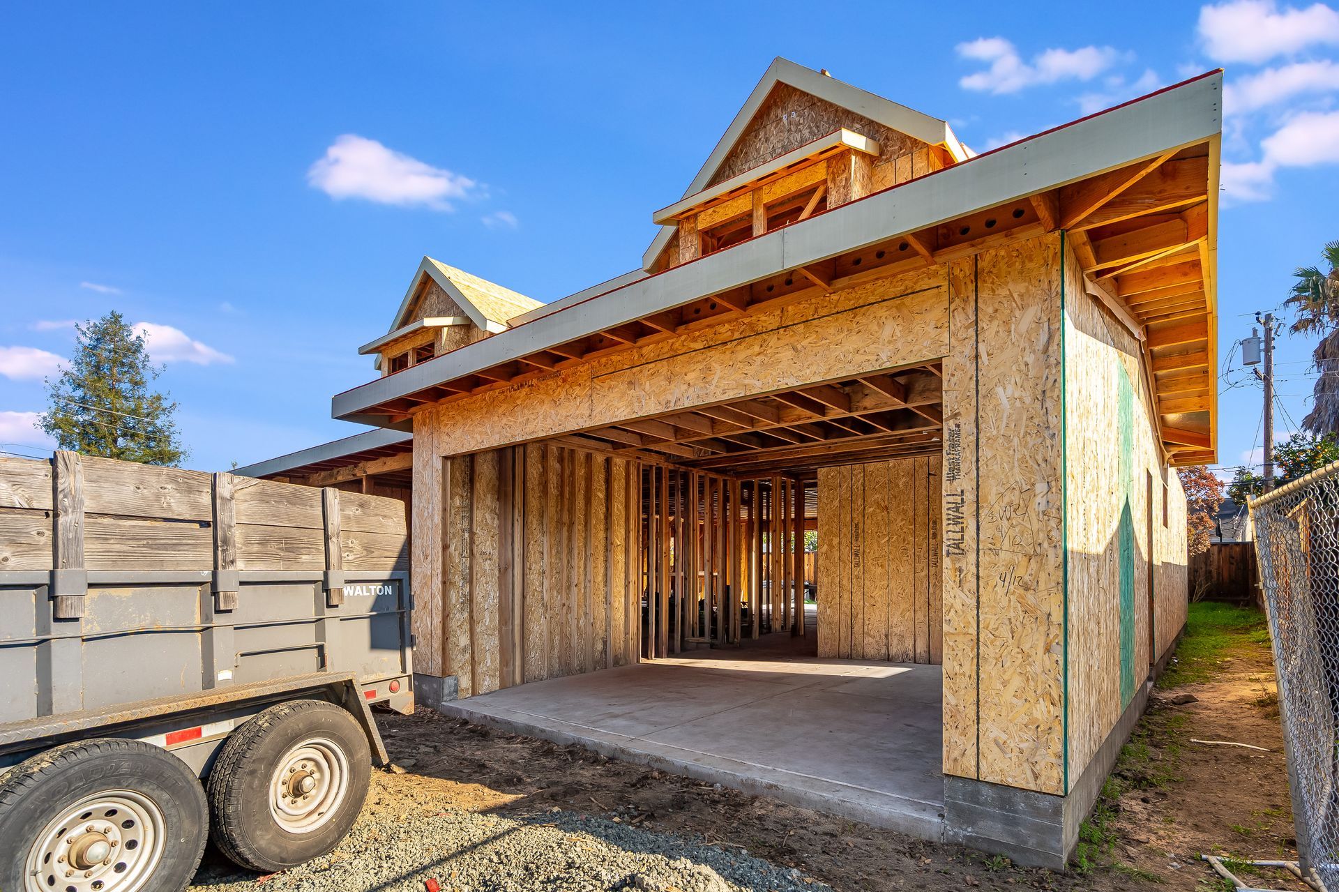 Unfinished house with open garage. Wooden frame construction; grey trailer parked in the foreground, blue sky background.