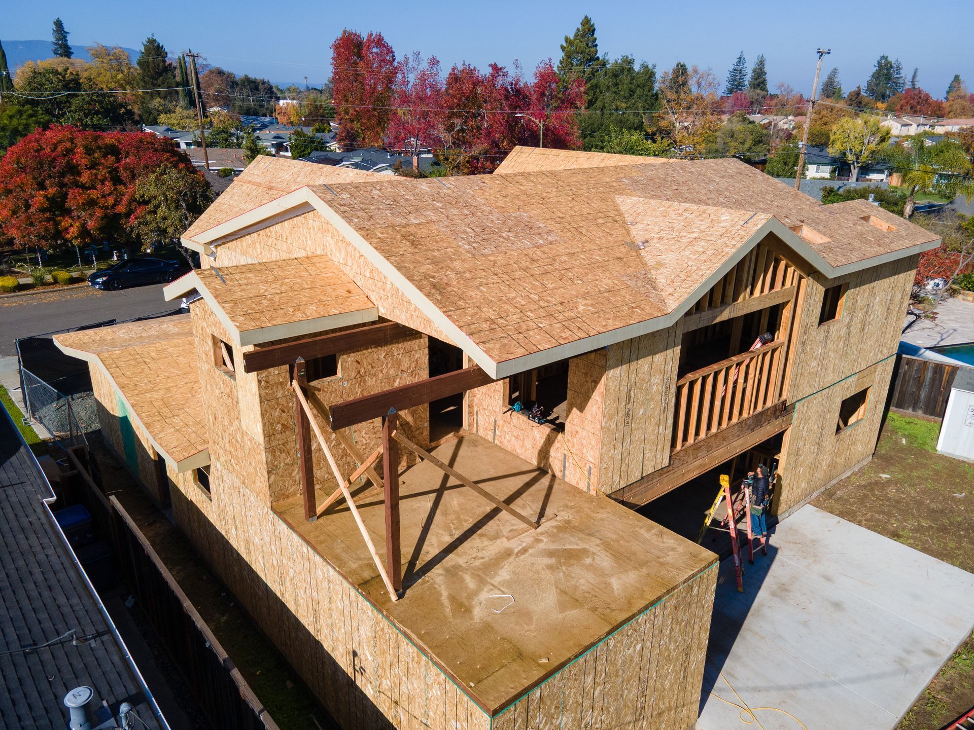 A house under construction with exposed wooden framework, people on the lower level, and fall foliage in the background.