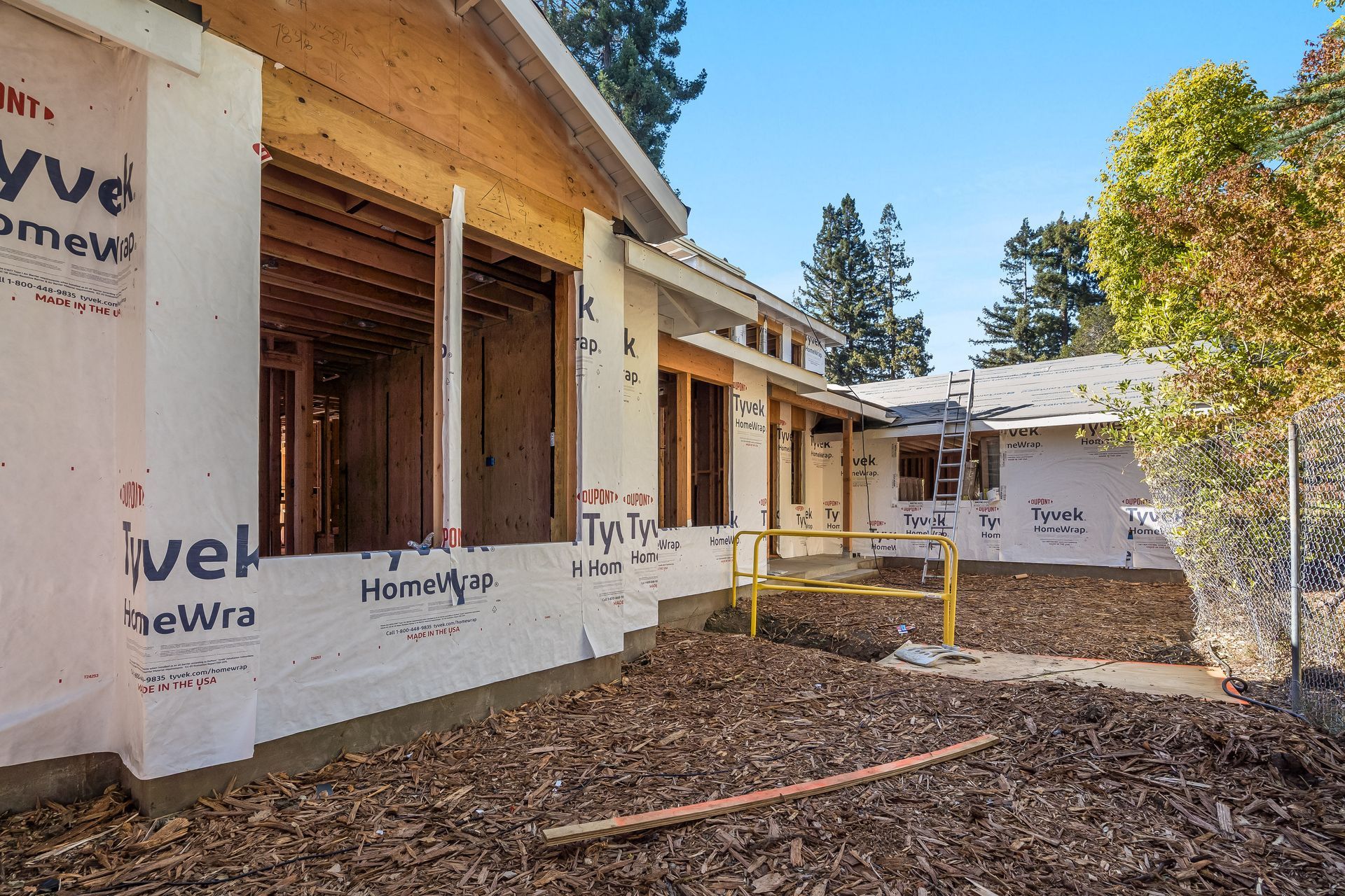 Building exterior under construction with exposed wood framing, Tyvek wrap, and debris.