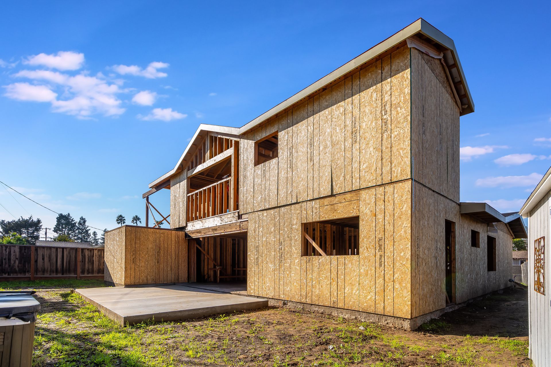 Two-story house under construction, with exposed wooden frame and sheathing against a blue sky.