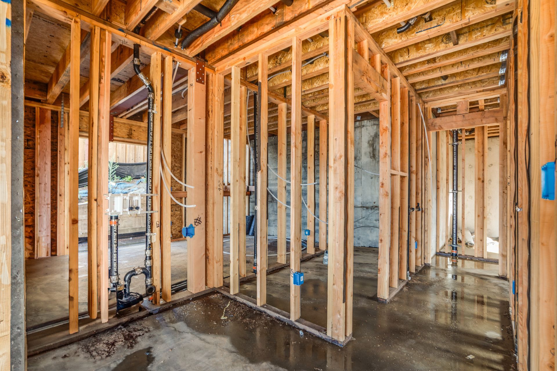 Interior view of a building under construction, wooden framework, concrete floor, visible plumbing and electrical work.