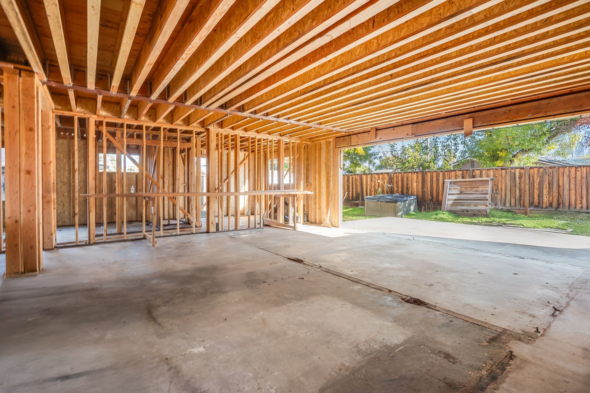 Interior of a building under construction, showing wooden framework and concrete floor, with a view of a backyard.
