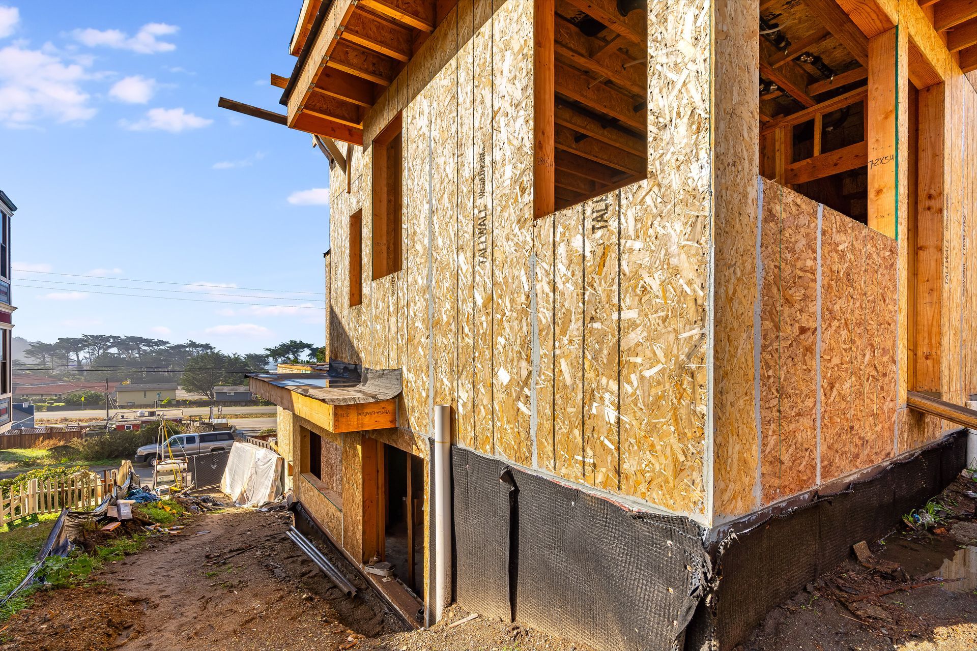 House under construction; wooden frame and roof trusses on dirt, blue sky.