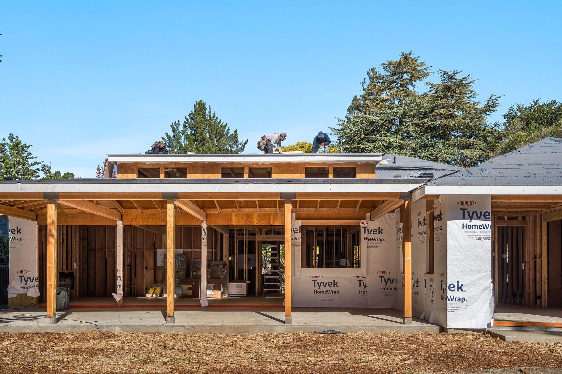 Construction site: Workers on roof, framing, Tyvek wrap, porch, blue sky.