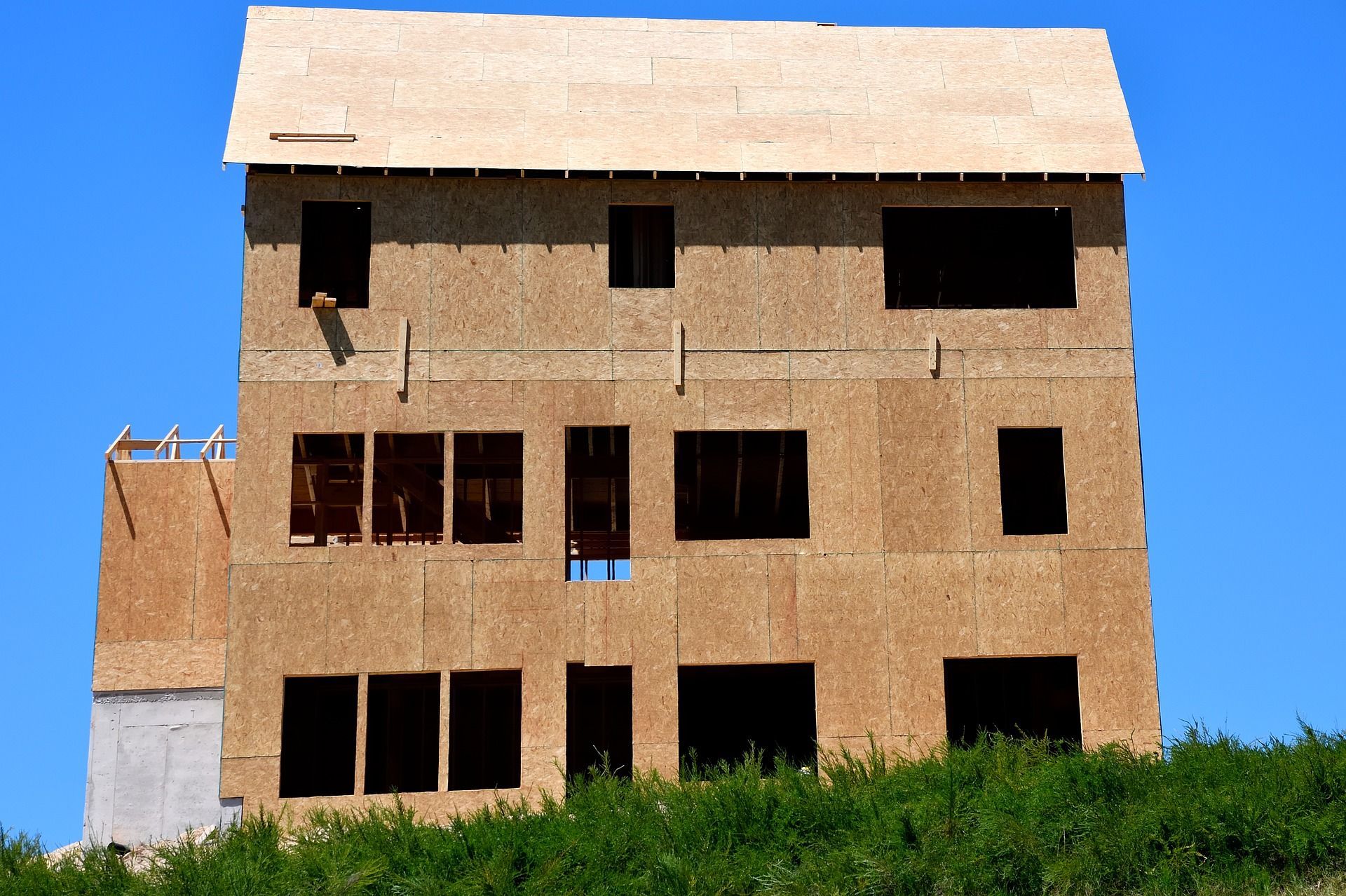 Unfinished two-story house with plywood siding and an exposed roof frame, against a blue sky.