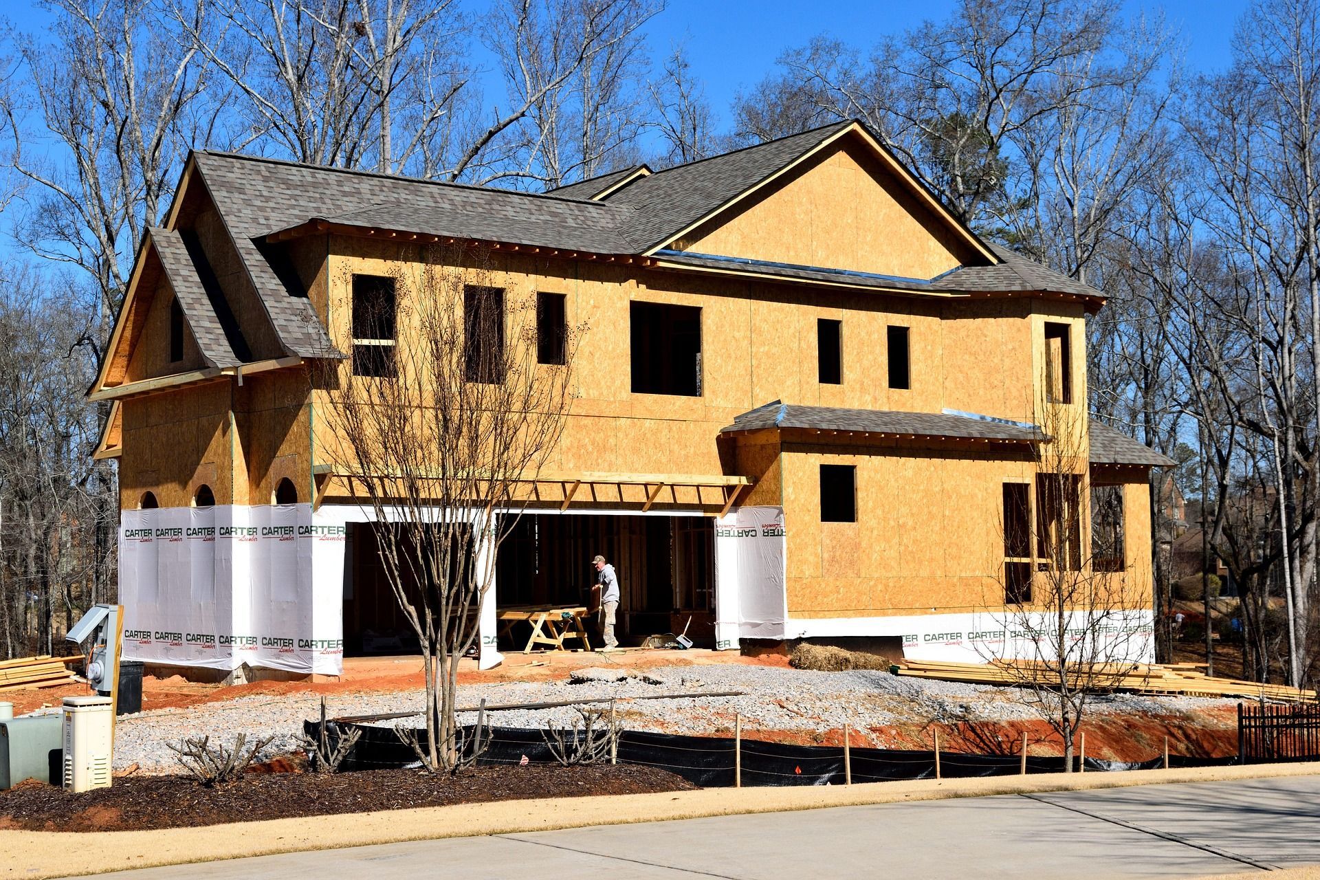 Two-story house under construction, with exposed wooden frame. A worker stands inside the carport.