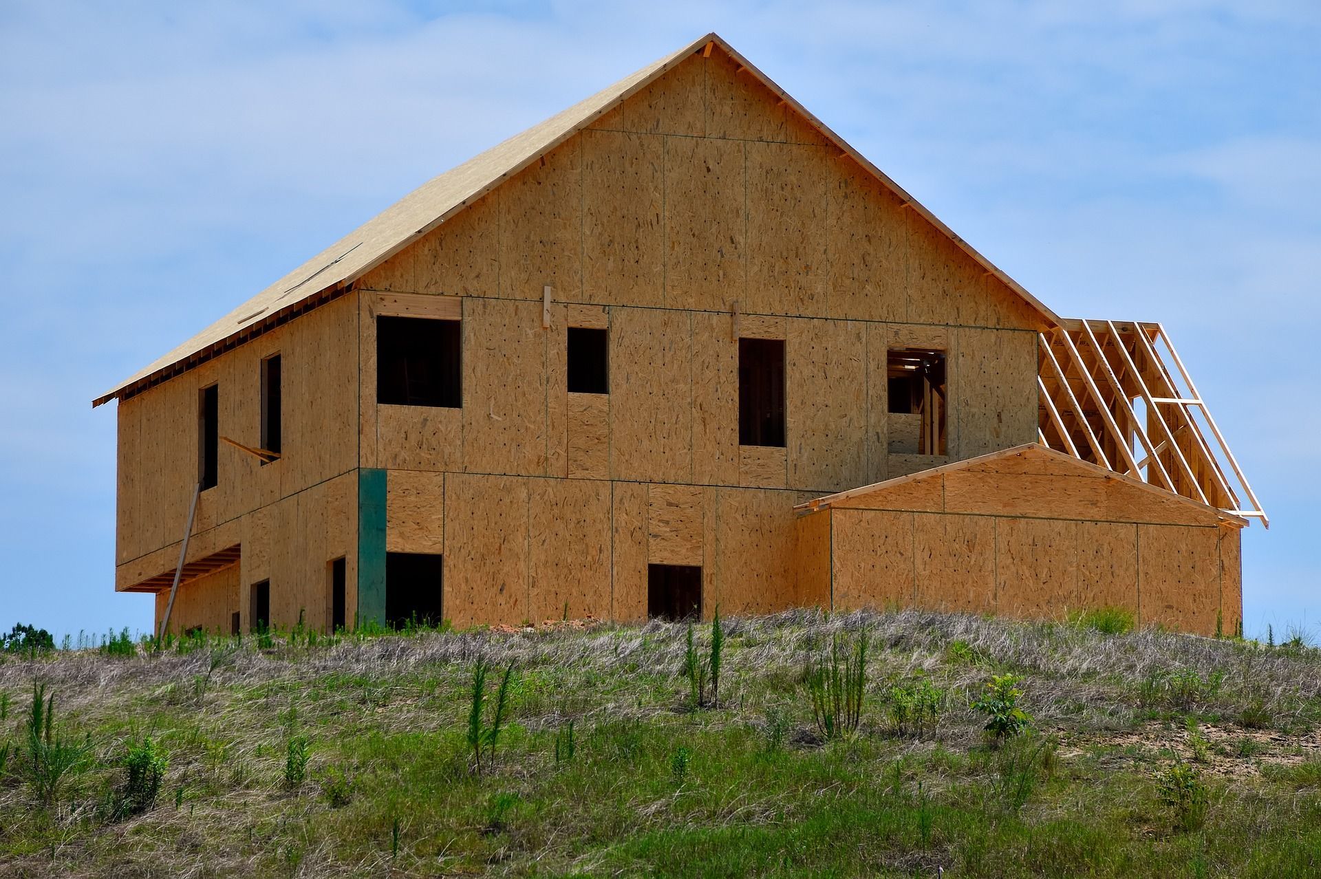 House under construction, wood-paneled walls, roof framed, set on a grassy hill, blue sky.