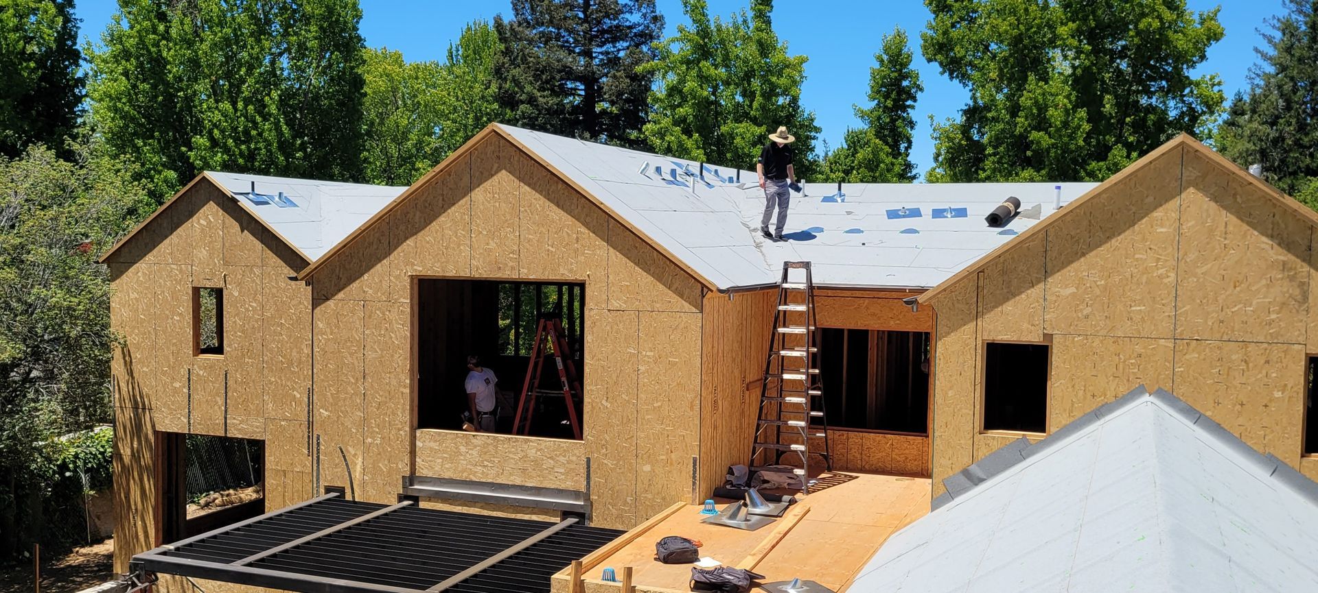 House under construction, workers on the roof, blue sky, trees in the background.