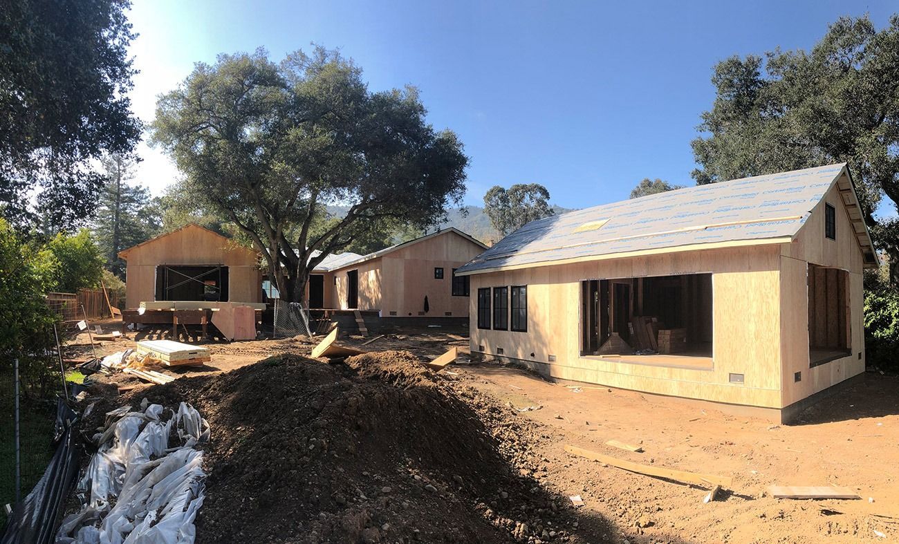 Construction site with three wooden buildings under construction, surrounded by dirt and trees.
