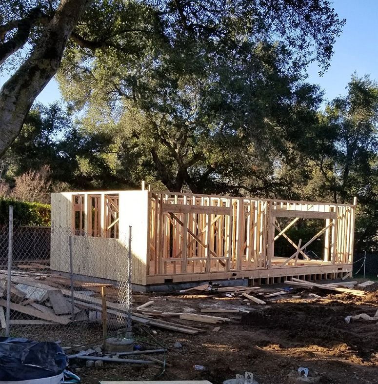 Wooden framework of a building under construction, outdoors with wood debris and trees in the background.