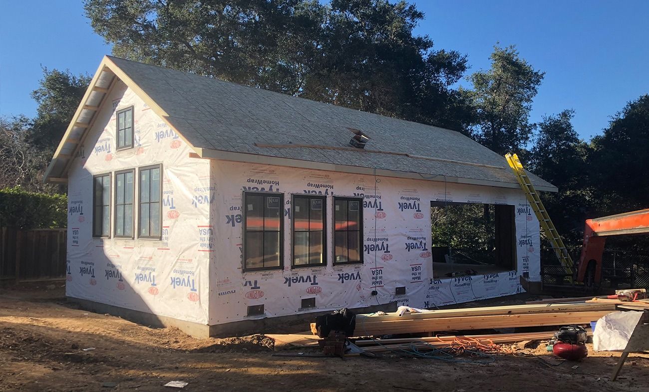 Construction of a small house, exterior view. White paper siding, black window frames, and wood beams.
