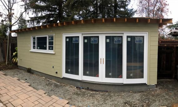Green shed with sliding glass doors and small window, gravel base, and brick walkway.