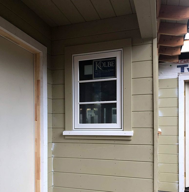 White-framed window on a building with olive siding. Kolbe logo visible on window. Construction in progress.