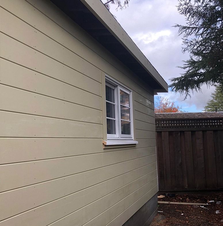 Beige building side with a white-framed window. Brown fence and cloudy sky in the background.