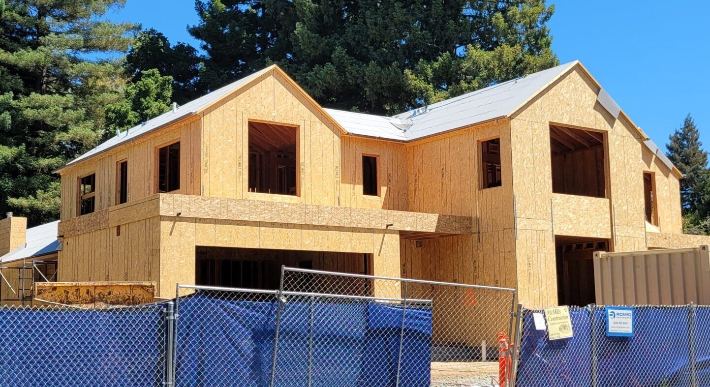 Unfinished two-story house under construction, framed with plywood, behind a blue chain-link fence.