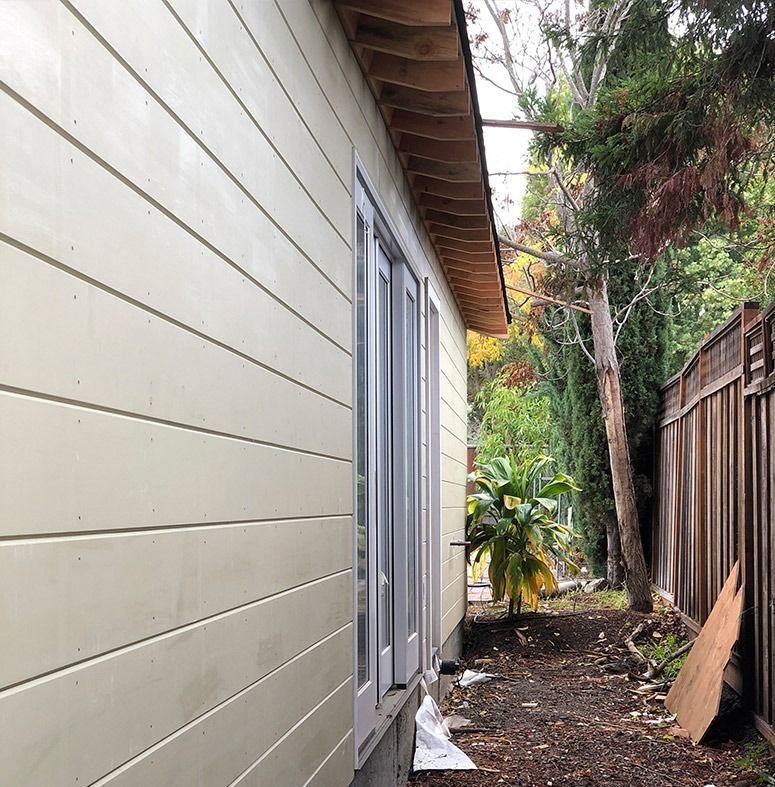 Side view of a building with light-colored horizontal siding, a window, and a narrow dirt path next to a fence.