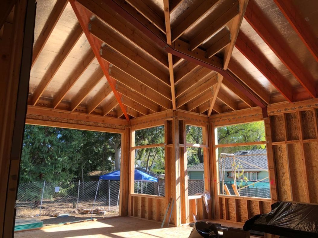 Interior view of a wooden building frame with angled ceiling and window openings, sunlight streaming through.