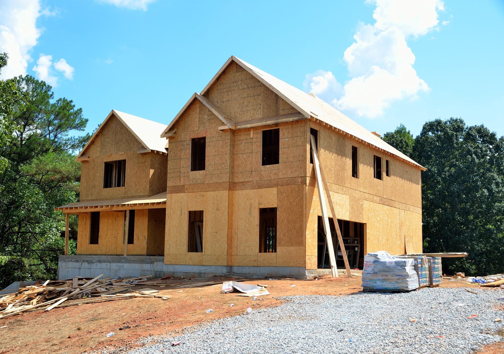 Two-story house under construction, with wood siding and open window frames, in a dirt and gravel lot under a blue sky.