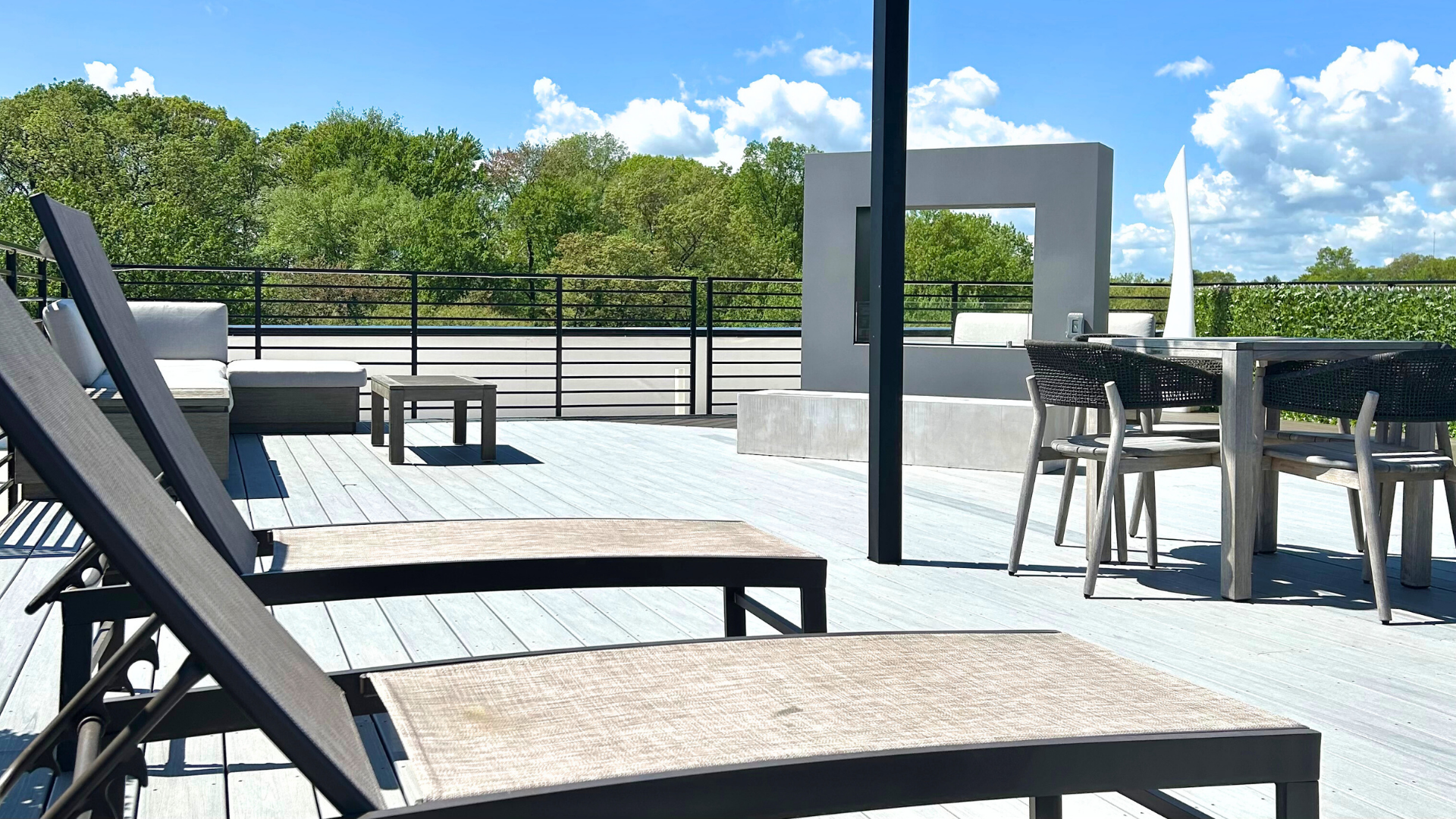 Modern rooftop lounge chairs with fireplace, lush green tree line view, and blue sky with white clouds