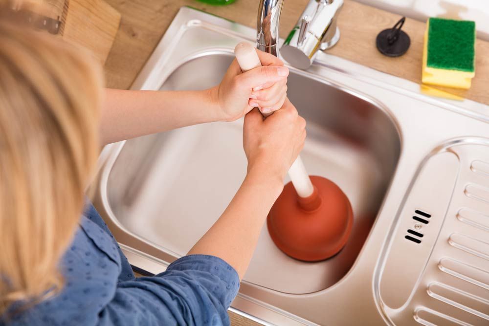 A Woman is Using a Plunger to Unblock a Sink — Absolute Plumbing Australia in Darwin, NT