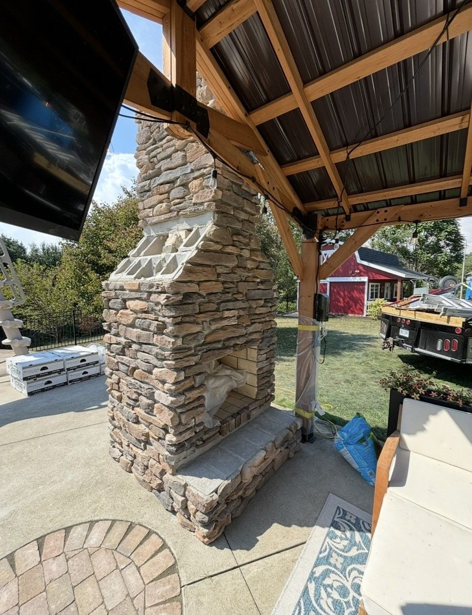 A stone fireplace is under a wooden canopy on a patio.