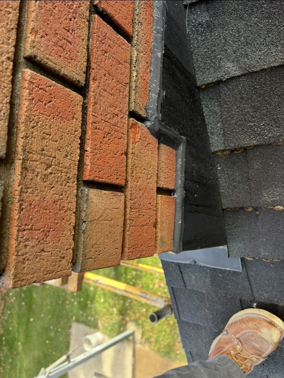 A close-up view of a brick wall intersecting with a shingled roof, showing black flashing and part of a brown work boot.