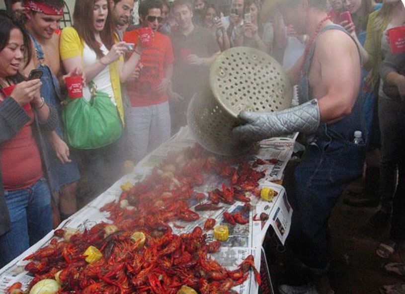 Man emptying a large basket of cooked crawfish onto a newspaper-covered table; crowd watches expectantly.