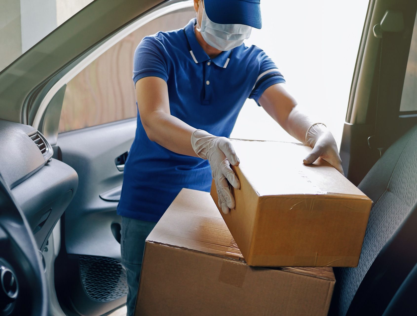 A delivery man wearing a mask and gloves is loading boxes into a car.