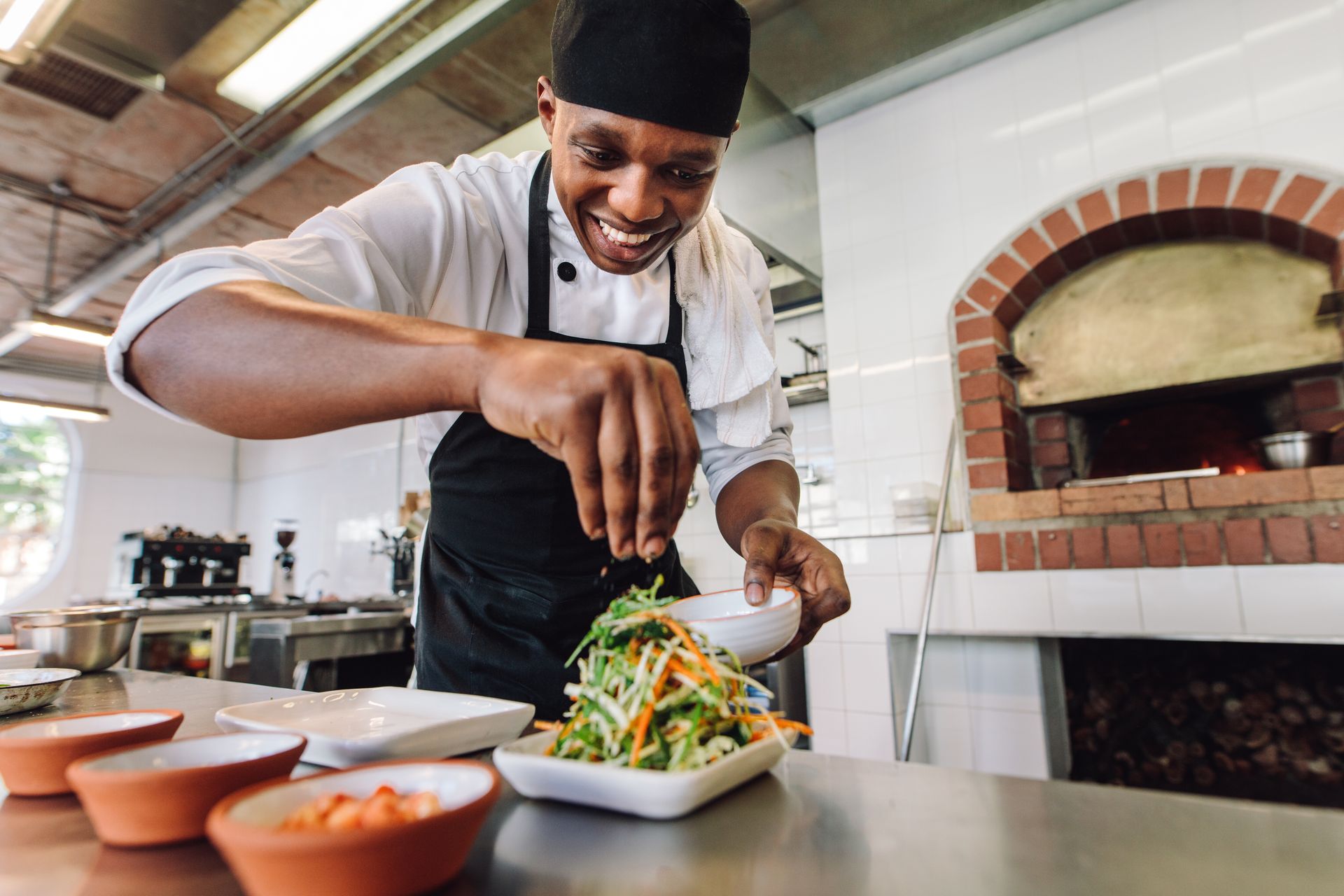 Chef smiles while seasoning salad in a restaurant kitchen, near a brick oven.