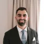 Portrait of Ameer Rebehy with a beard is wearing a suit and tie and smiling .