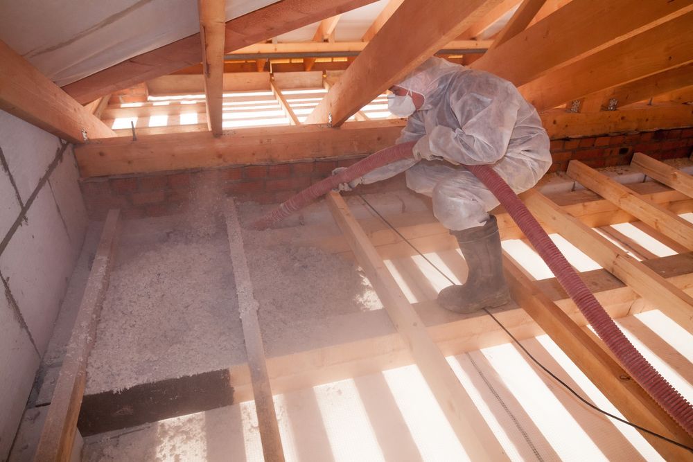 Worker in white coveralls on attic beams, spraying insulation in a wooden roof space.