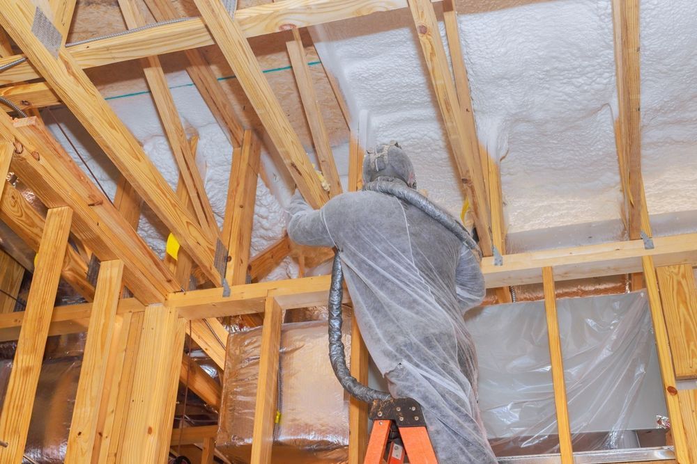 Worker spraying insulation foam between exposed wooden roof beams in an unfinished attic