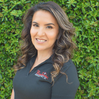 A woman in a black shirt is smiling in front of a hedge.