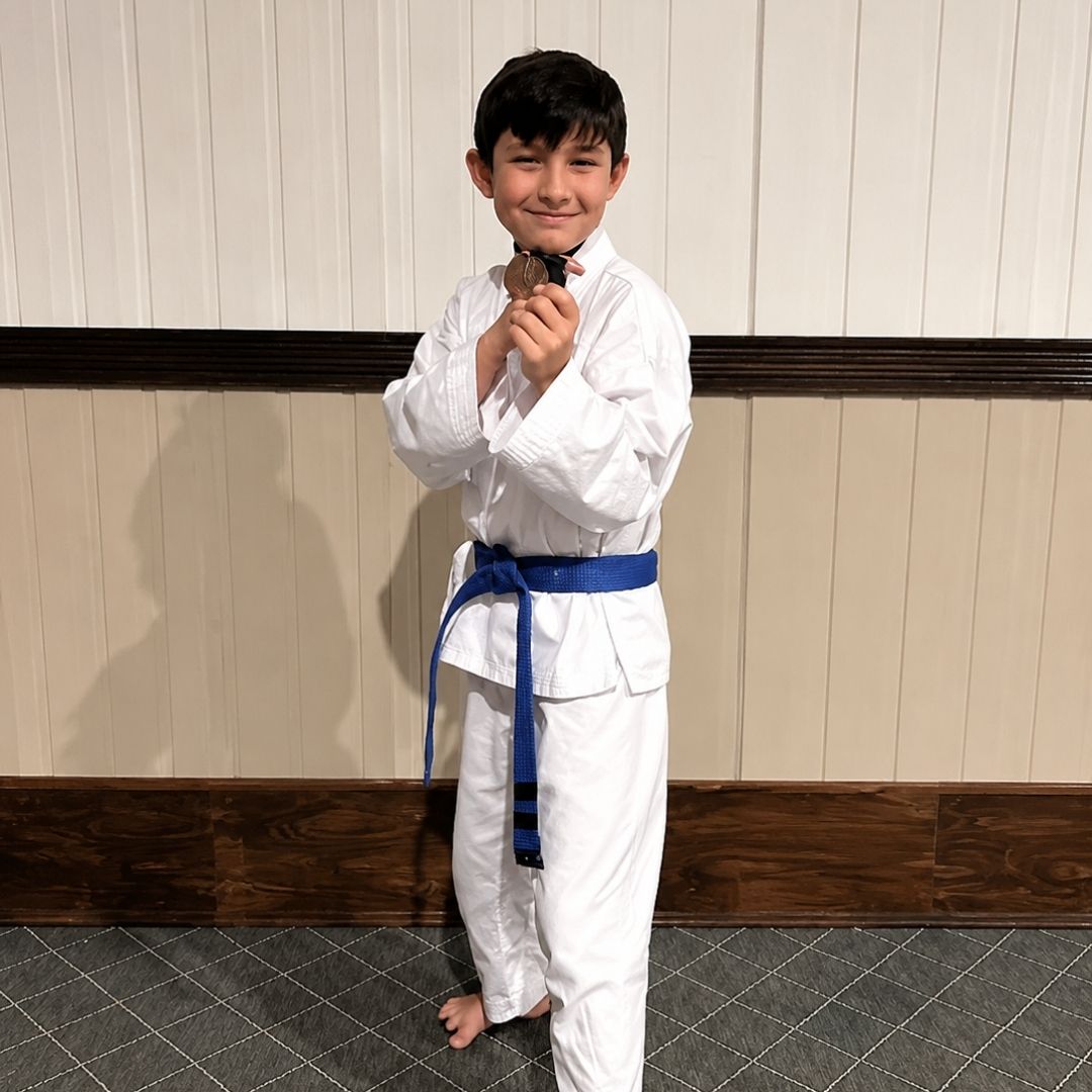 Two children practicing Brazilian Jiu-Jitsu on a mat. One child in white gi has the other in a leg lock. A girl watches.