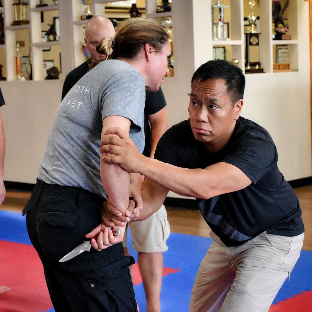 Five people in jiu-jitsu gear pose together in a gym. Smiling, some have arms around each other. Machado logo visible.