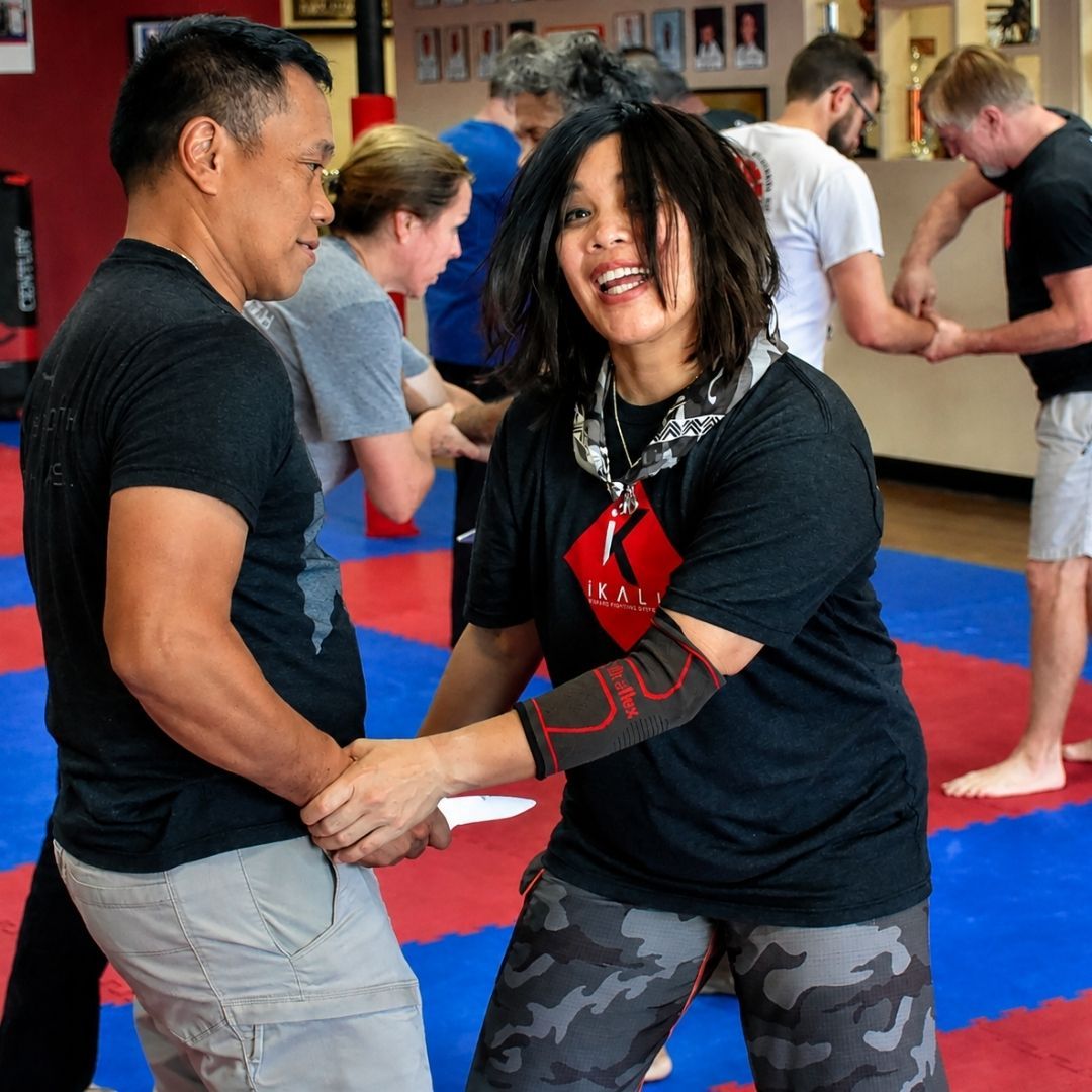 Two men in grappling position on a mat. One embraces the other from behind, both smiling. Gym setting.