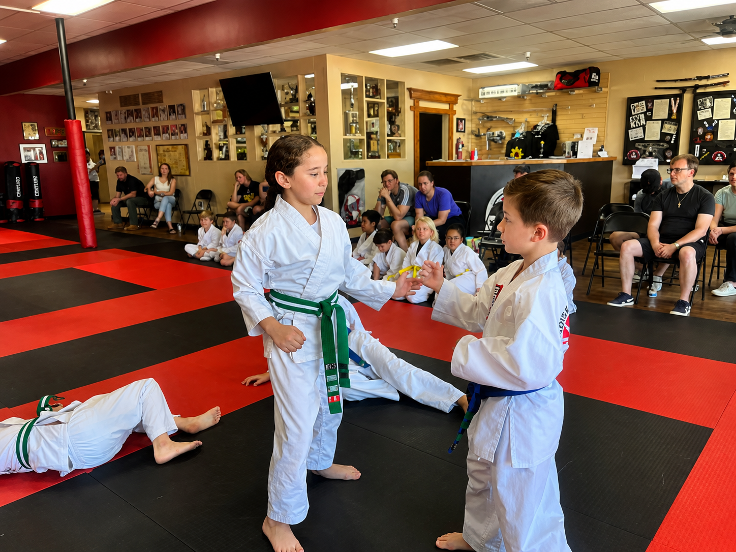 Five people in jiu-jitsu gear pose together in a gym. Smiling, some have arms around each other. Machado logo visible.