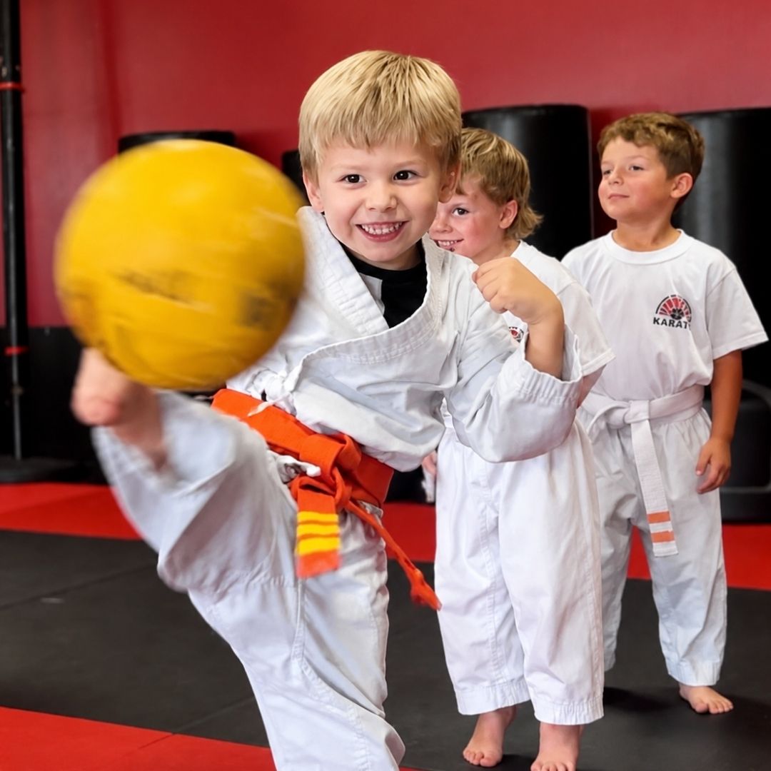 Two children in black and white martial arts uniforms practice a chokehold on a mat. One child smiles.