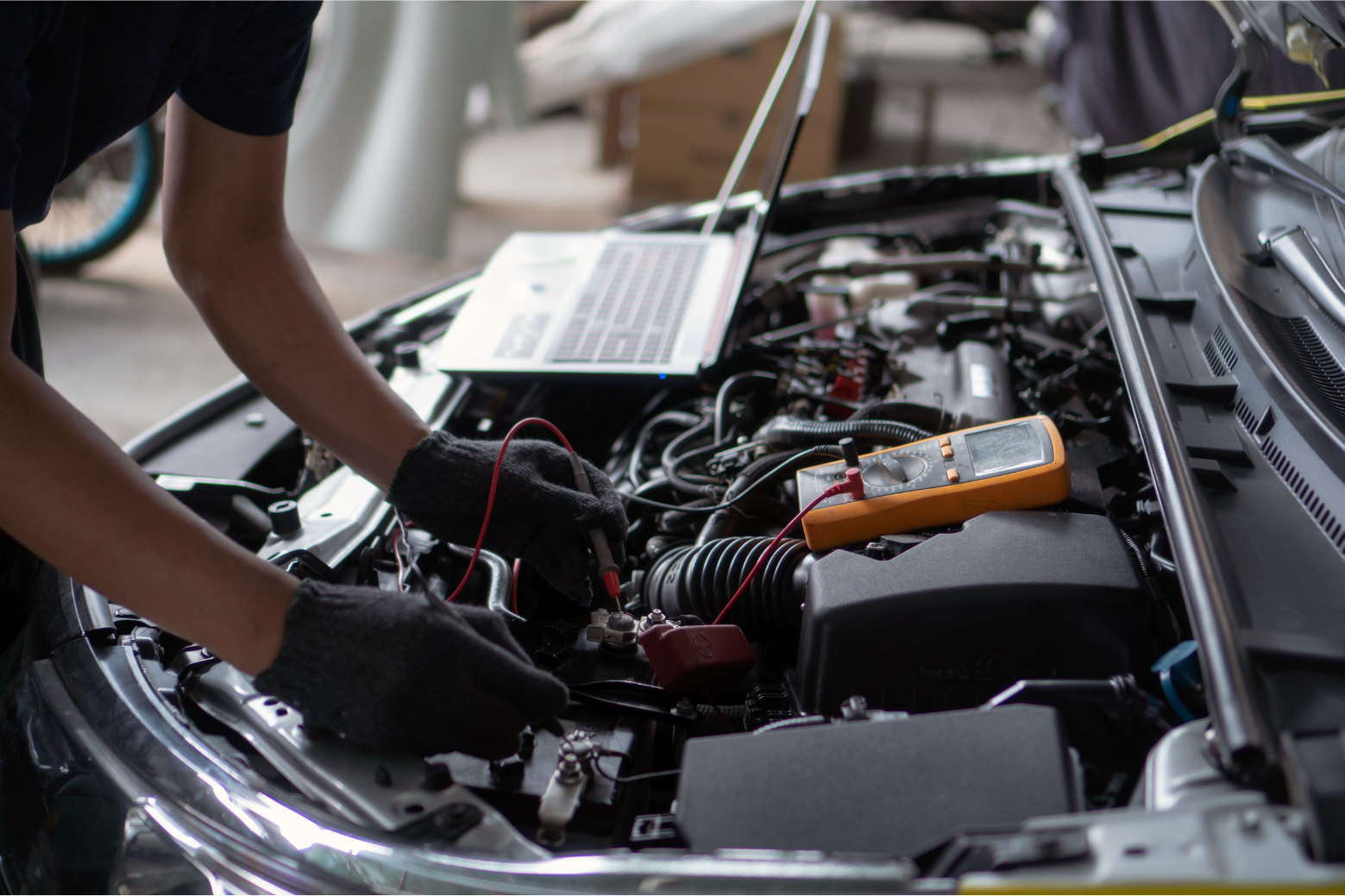A man is working on a car engine with a laptop and a multimeter.