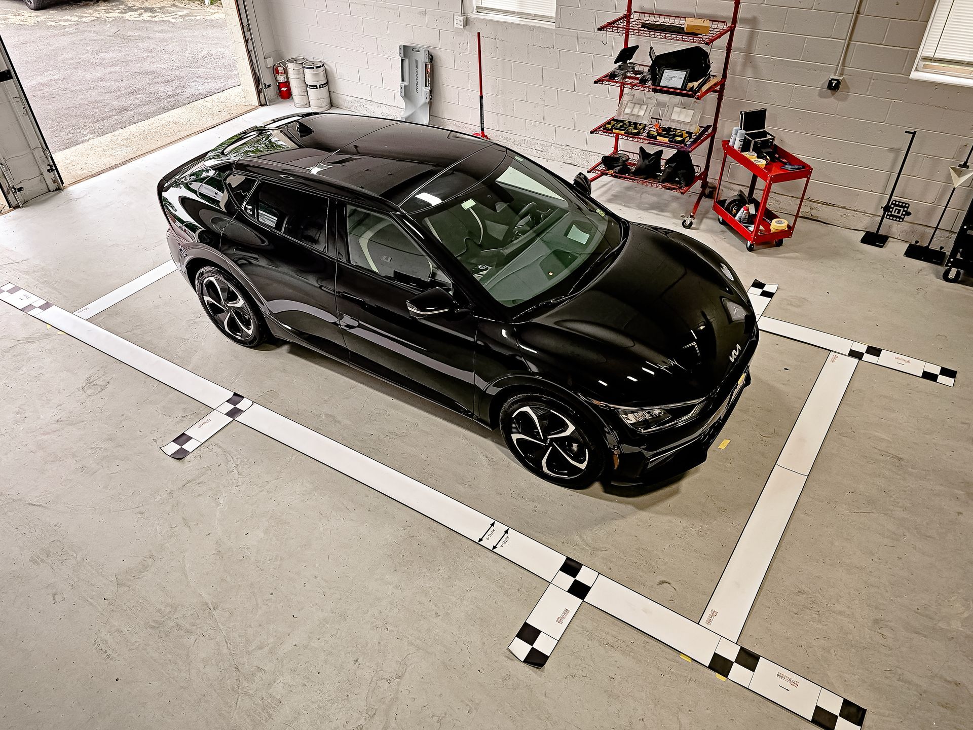 A black car is parked in a garage with ADAS calibration targets on the floor.