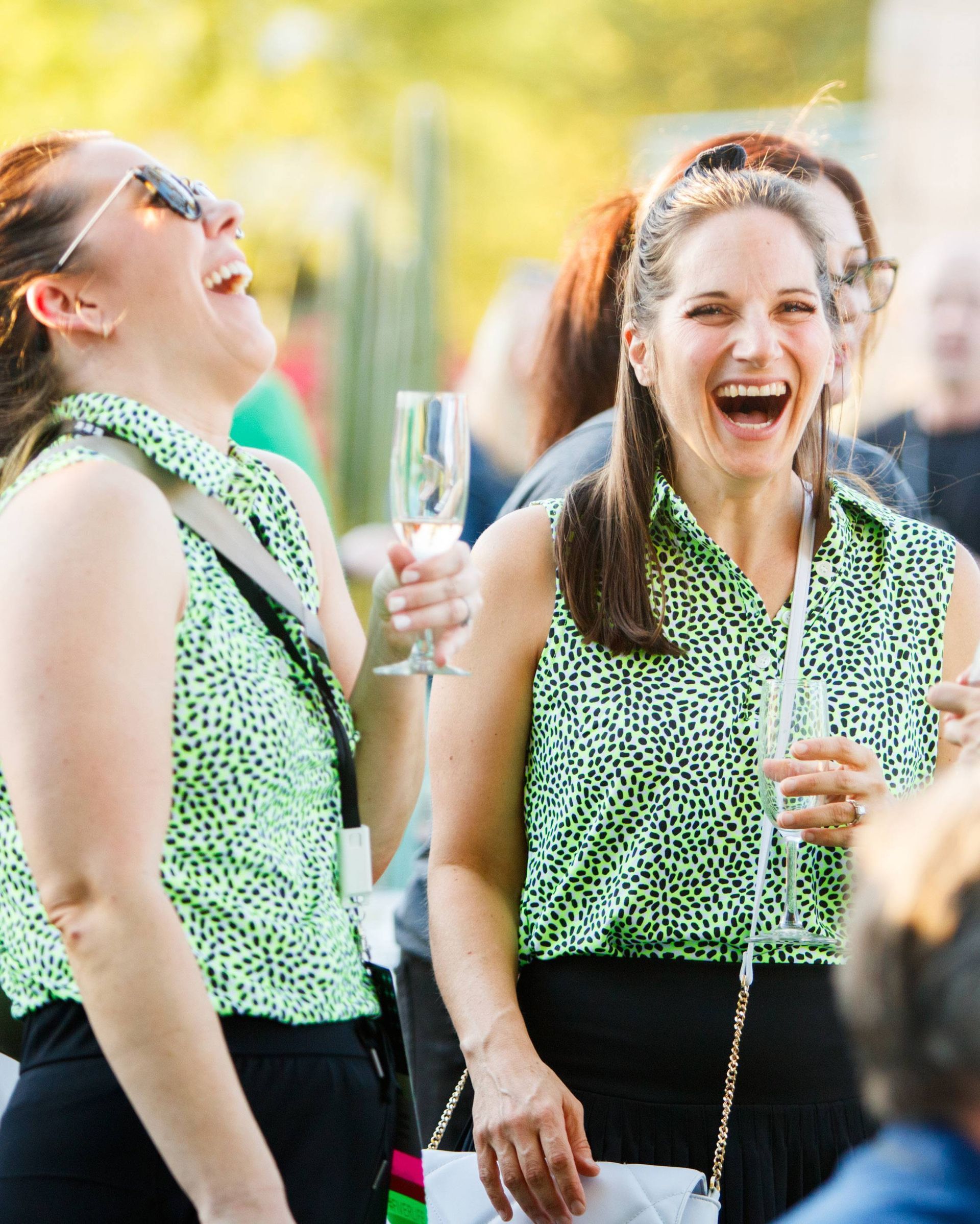 Two women in green shirts are laughing and holding wine glasses