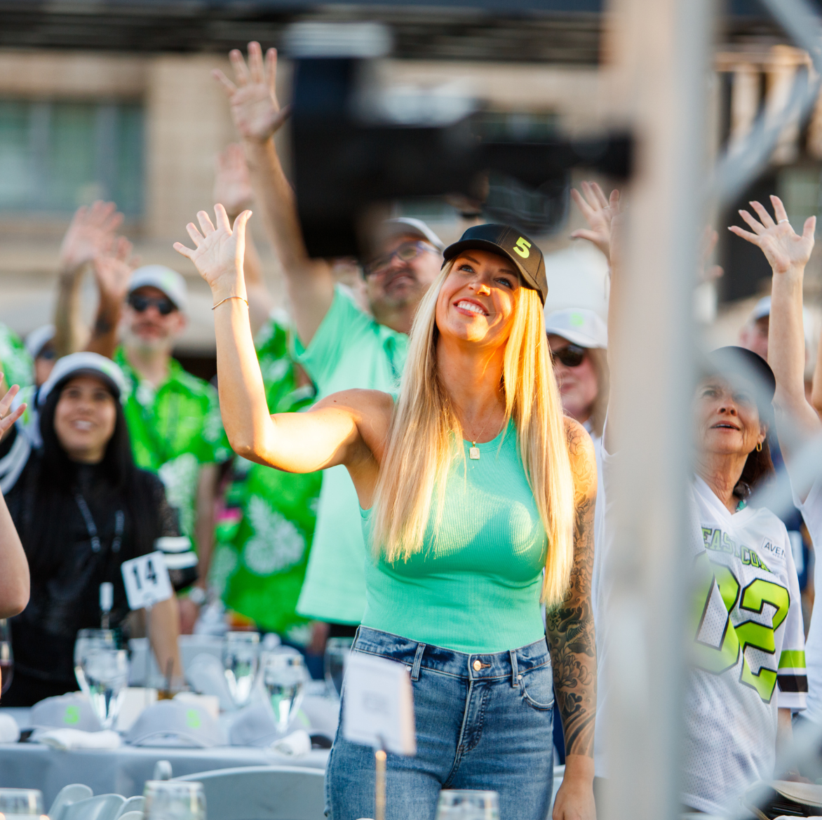 A woman in a green tank top stands in front of a crowd of people and waves.