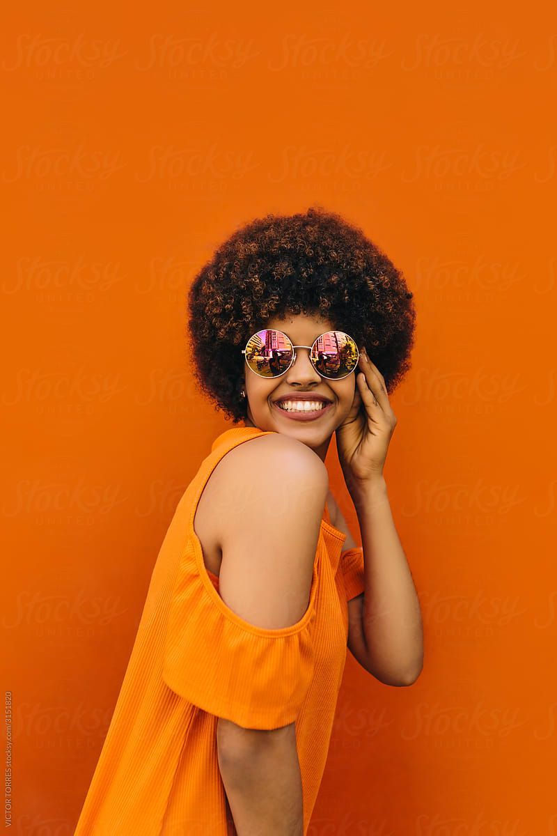 A woman wearing sunglasses with flowers on them is smiling in front of an orange wall.