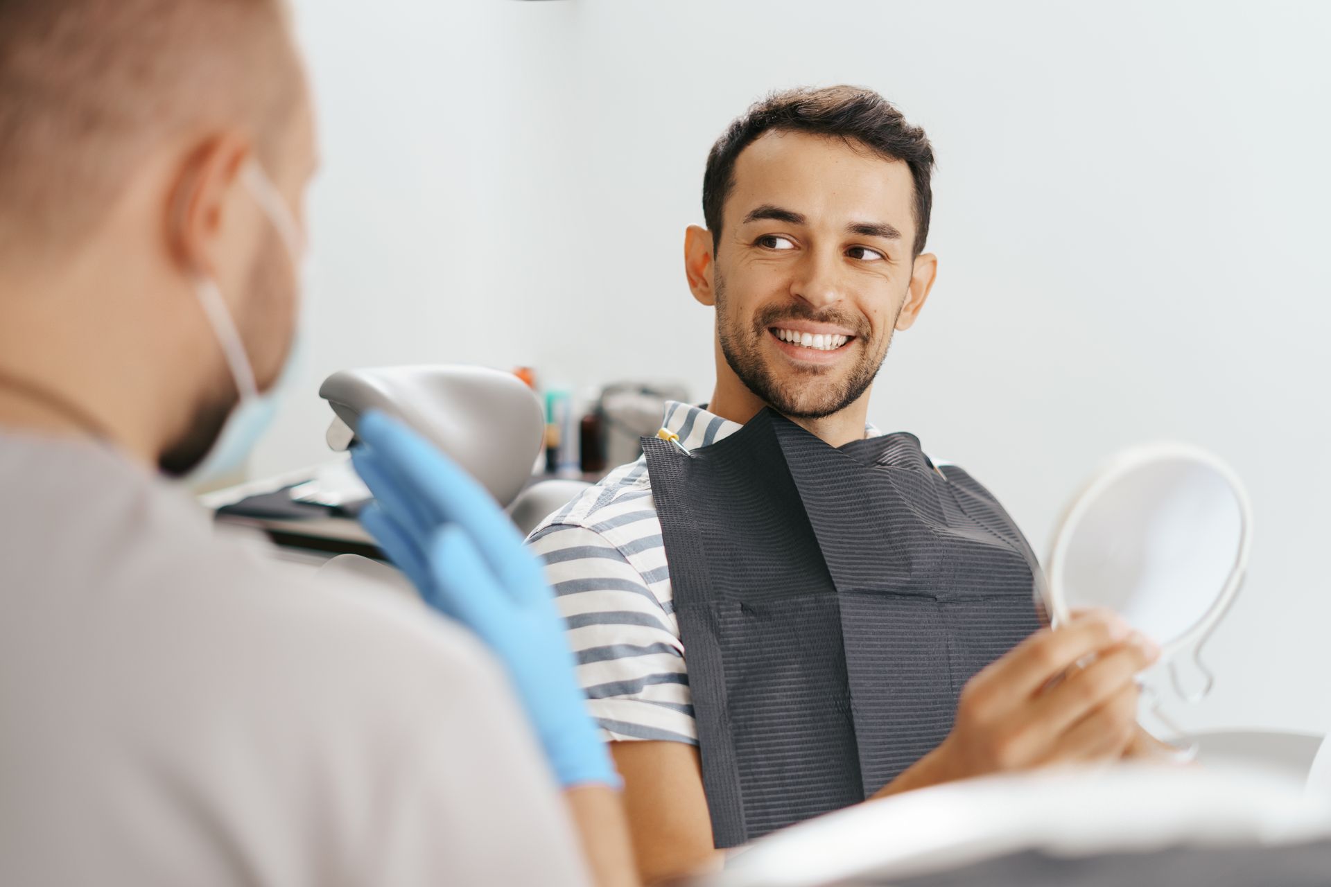 A man is sitting in a dental chair looking at his teeth in a mirror.