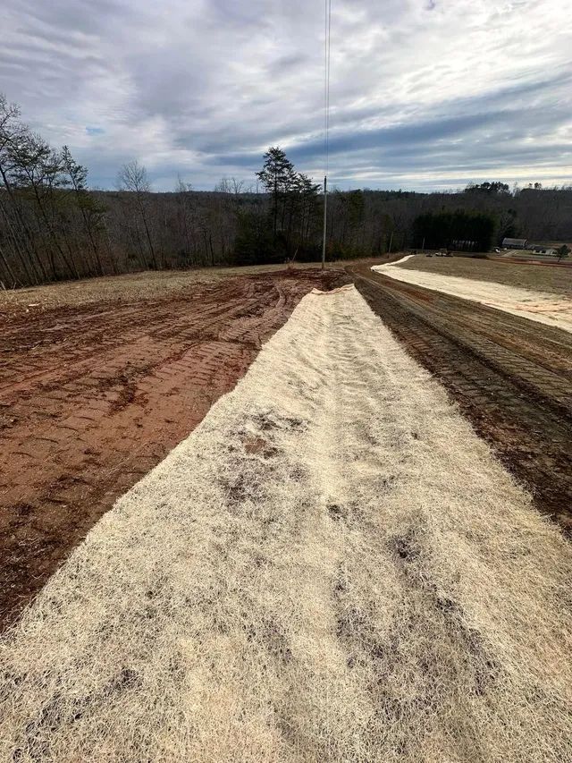 A dirt field featuring a long strip of straw erosion control matting, with a pole standing in the background.