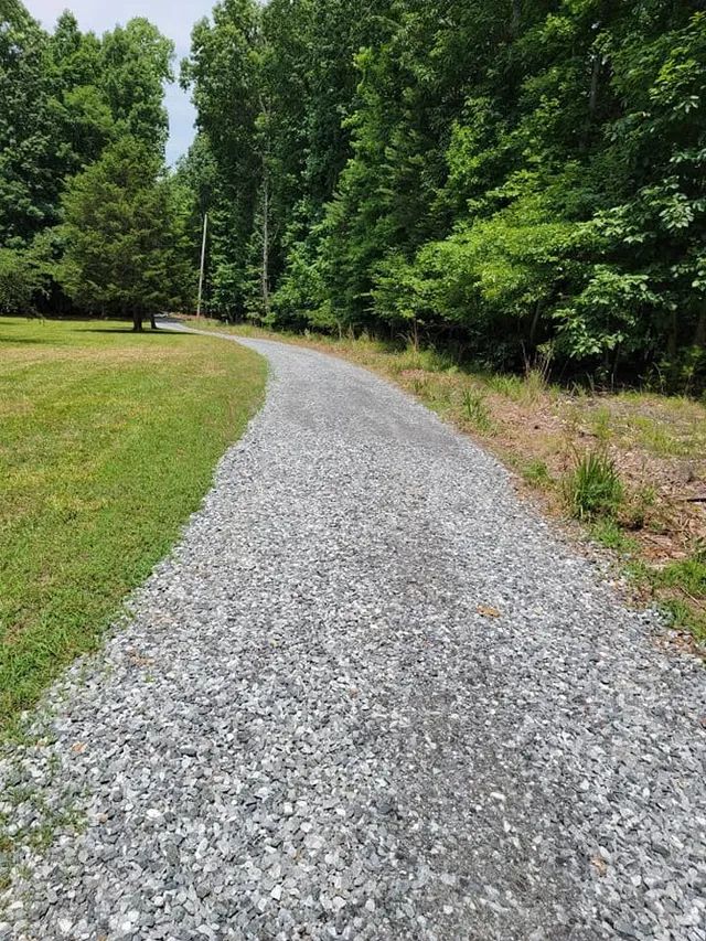 A gravel path curves through a grassy clearing bordered by a dense forest under a bright sky.
